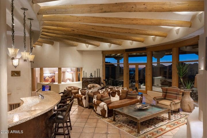 The soaring beamed ceilings and open-concept layout of this great room highlight sweeping mountain views and rich architectural design. Custom lighting and grand windows create a warm, inviting ambiance perfect for desert evenings in this hilltop luxury home.