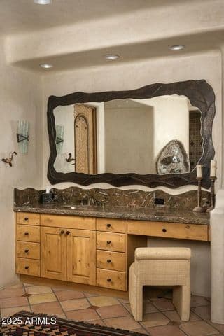 Showcasing two separate vanities, each with intricate stonework and hand-carved details, this expansive primary bathroom offers privacy and style. Warm wood cabinetry and desert-inspired tile work reflect the home’s luxurious Southwestern architecture.